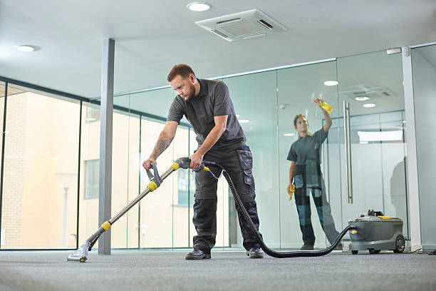 a male cleaning contractor steam cleans an office carpet in a empty office in between tenants. In the background a female colleague is polishing the glass partition offices .
